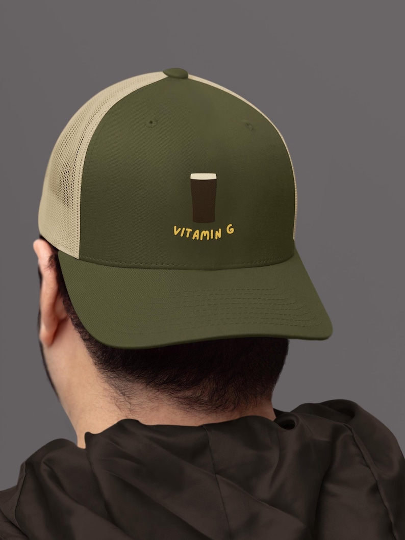 May include: Olive green trucker hat with a tan mesh back. The hat features a graphic of a dark brown pint of beer and the text "VITAMIN G" in yellow.