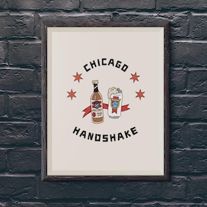 May include: A framed print with a white background and black text that reads "CHICAGO HANDSHAKE". The print features a cartoon illustration of a bottle of Mal&ouml;rt liquor and a can of beer with red, white, and blue stars.