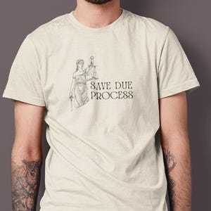 May include: A cream-coloured t-shirt with a black illustration of Lady Justice holding scales and a sword, and the text "SAVE DUE PROCESS". The shirt is made of a soft material.