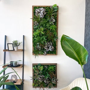 Op de afbeelding: Twee ingelijste verticale groene wandkunstwerken met een verscheidenheid aan planten en bladeren. Het bovenste stuk is rechthoekig en het onderste stuk is vierkant. De wandkunst hangt aan een witte muur met een witte stoel en een groene plant op de voorgrond.