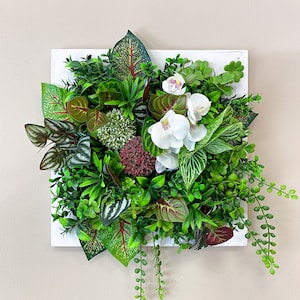Op de afbeelding: Een wit vierkant frame met een groene en witte bloemschikking. De schikking omvat een ​​verscheidenheid aan groene planten, witte orchideeën en een paar rood getinte bladeren.