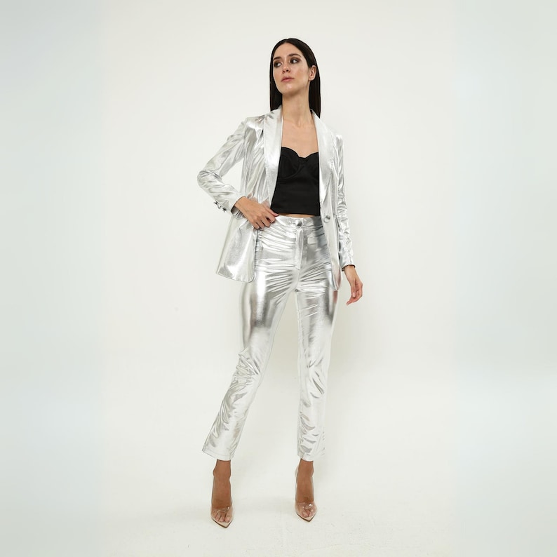 May include: A silver metallic blazer and pants suit. The blazer has a notched lapel and long sleeves. The pants are slim-fitting and have a high waist.