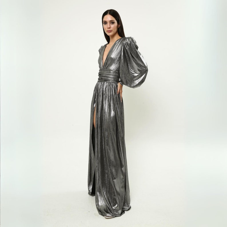 May include: A silver metallic maxi dress with long sleeves and a V-neckline. The dress has a fitted bodice and a flowing skirt with a thigh-high slit on each side.