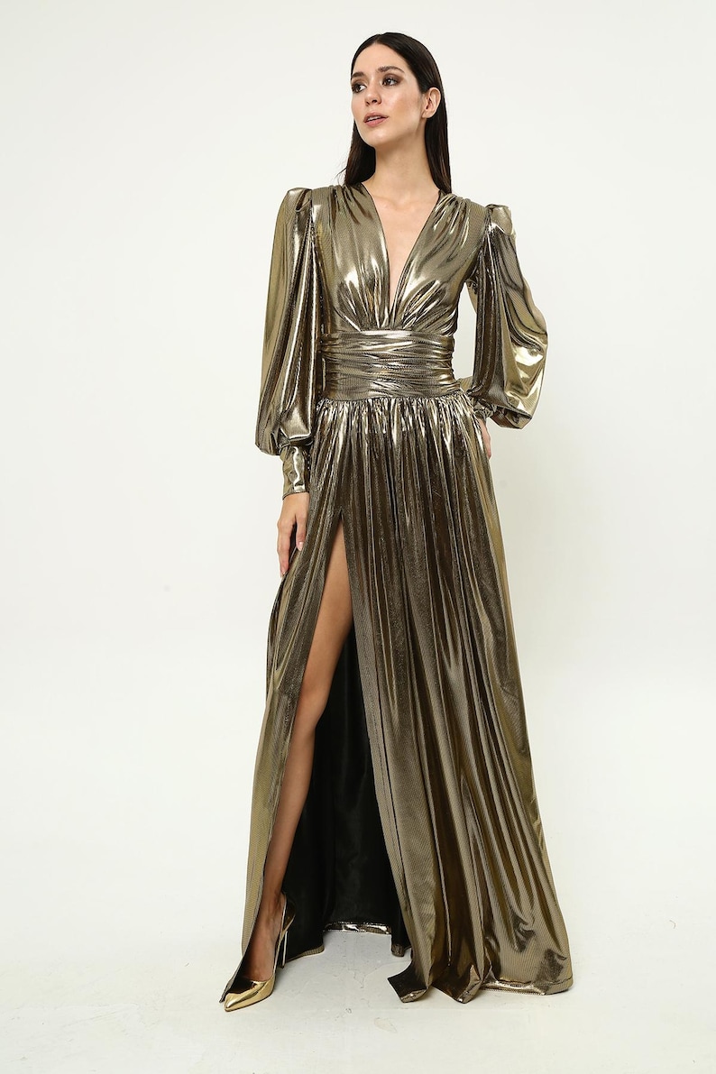 May include: A long, gold metallic dress with a plunging neckline, long sleeves, and a thigh-high slit. The dress has a fitted bodice and a flowing skirt.