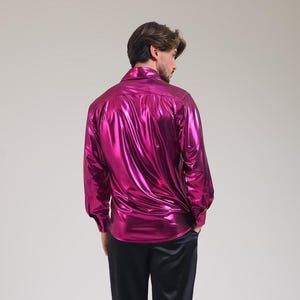 K&ouml;nnte beinhalten: Ein gl&auml;nzendes, magentafarbenes Langarmhemd mit klassischem Kragen. Das Hemd hat ein gl&auml;nzendes, metallisches Finish und wird mit einer schwarzen Hose kombiniert. Der Stoff reflektiert das Licht und erzeugt einen lebendigen und auff&auml;lligen Look.