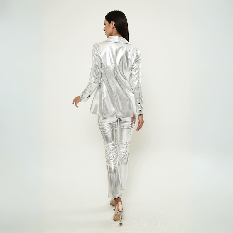 May include: A silver metallic blazer and pants suit. The blazer has a notched collar and long sleeves. The pants are slim-fitting and have a straight leg.