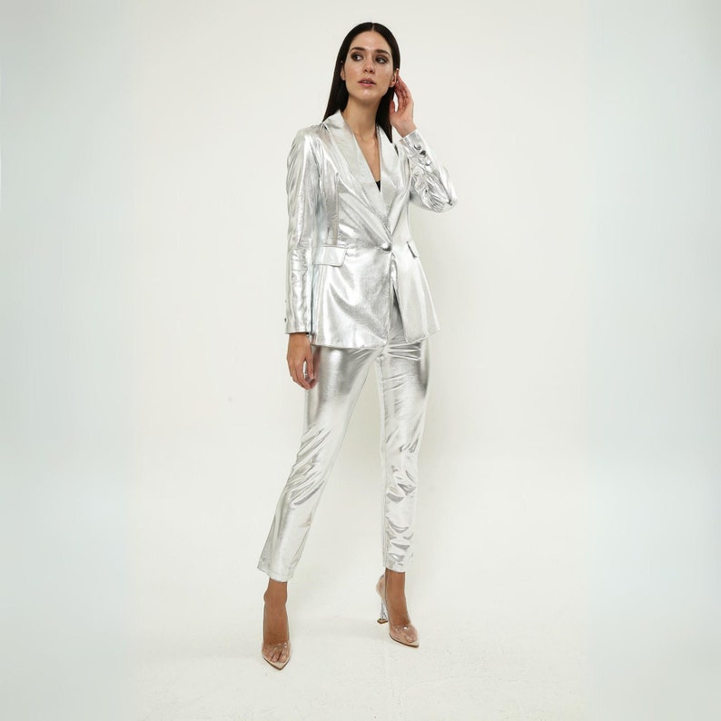 May include: A silver metallic blazer and pants suit. The blazer has a notched lapel and a single button closure. The pants have a straight leg.