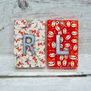 May include: Two resin keychains with silver letters "R" and "L" embedded in the resin. The keychain with the letter "R" has a white background with a pattern of pink and white cartoon characters. The keychain with the letter "L" has a red background with a pattern of pink and white cartoon characters.