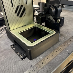 May include: A black and silver industrial machine with a hinged top and a rectangular base. A ruler with inch markings is visible. The machine has a circular vent and a sliding tray. A complex mechanical component is attached to the side.