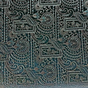 Peut inclure: Gros plan sur un tissu bleu canard avec un motif complexe de broderies argentées et bleu canard. Le motif présente des formes florales et géométriques, notamment des feuilles stylisées, des cercles et des cadres rectangulaires. Le tissu a un aspect texturé.
