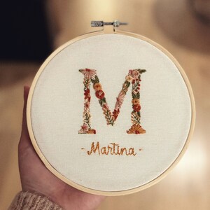 May include: Embroidered hoop art featuring the letter "M" made of colorful flowers and the name "Martina" written below.