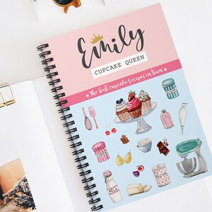 May include: A pink and blue spiral bound notebook with the title "Emily Cupcake Queen" and the text "The best cupcake recipes in town." The notebook features illustrations of cupcakes, baking ingredients, and kitchen tools.
