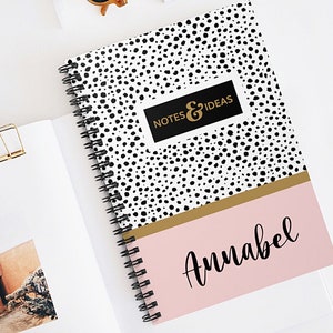 May include: A black and white polka dot notebook with a gold stripe and a pink section at the bottom. The notebook has a black and gold label that says "NOTES & IDEAS". The pink section has the name "Annabel" written in black cursive.