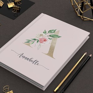 May include: A pink notebook with a gold letter A decorated with watercolor flowers and the name Annabelle. The notebook is on a brown surface with gold pencils and paper clips.