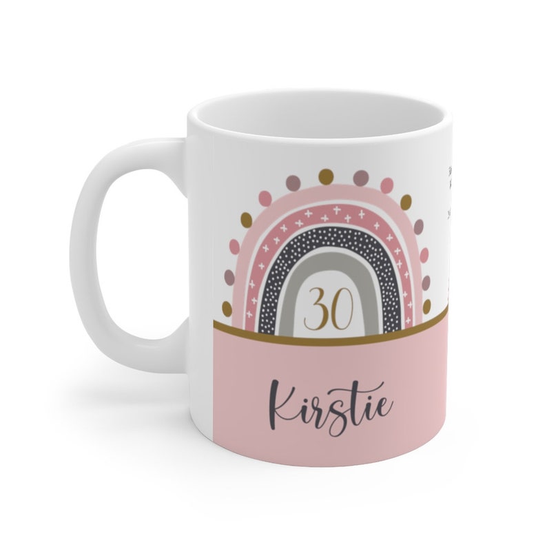Op de afbeelding: Witte keramische mok met een roze en grijze regenboogontwerp. De regenboog heeft gouden accenten en het getal 30 in gouden letters. De onderkant van de mok heeft een roze band met de naam Kirstie in gouden letters.