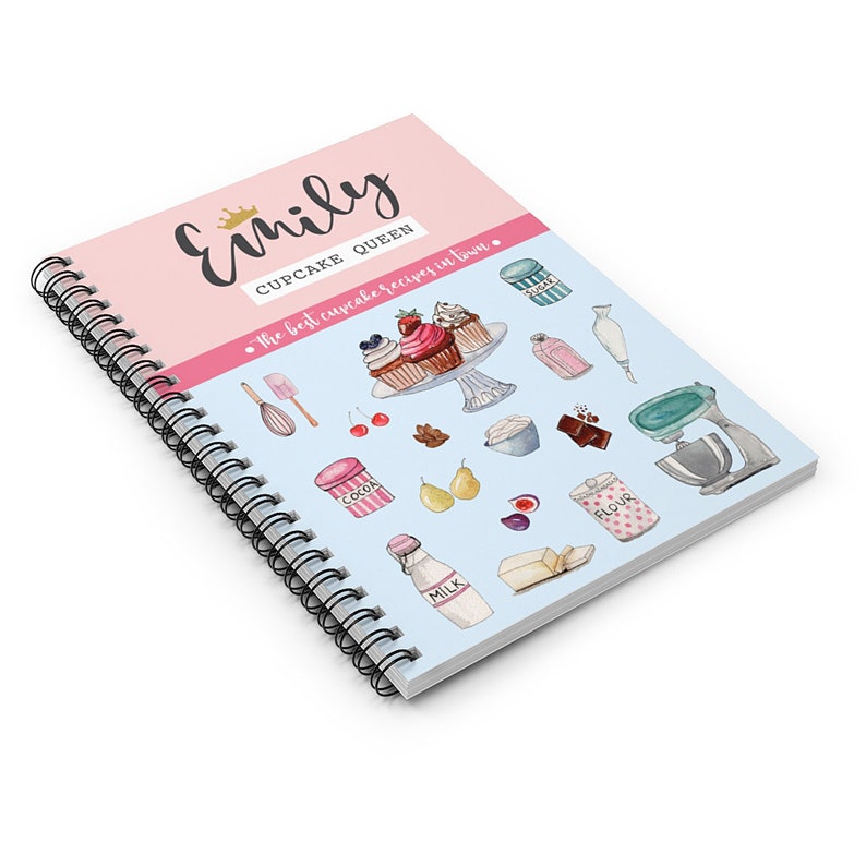May include: A pink and blue spiral bound notebook with the title "Emily Cupcake Queen" and the text "The best cupcake recipes in town." The cover features illustrations of cupcakes, baking ingredients, and kitchen tools.