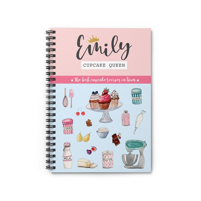 May include: A pink and blue spiral bound notebook with the title "Emily Cupcake Queen" and the text "The best cupcake recipes in town." The cover features illustrations of cupcakes, baking ingredients, and kitchen tools.