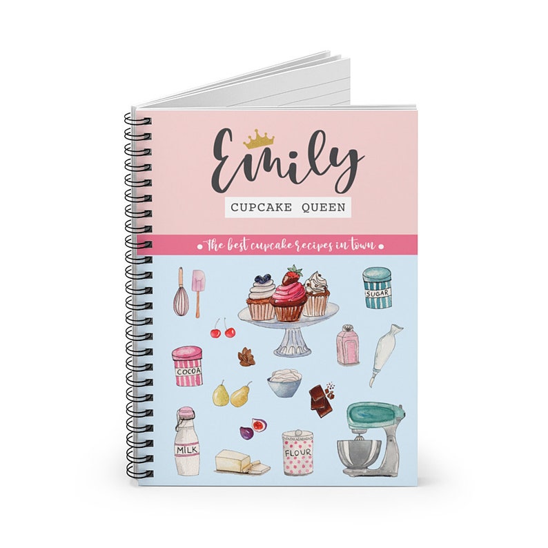 May include: A pink and blue spiral bound notebook with the text "Emily Cupcake Queen" and the words "The best cupcake recipes in town."  The cover features a watercolor illustration of cupcakes, baking ingredients, and kitchen tools.
