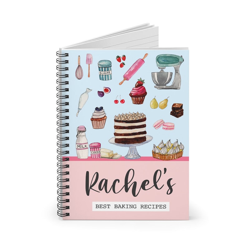 May include: A spiral bound notebook with a light blue cover featuring watercolor illustrations of baking ingredients and tools. The cover also has a pink stripe at the bottom with the text "Rachel's Best Baking Recipes".