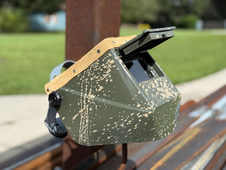 Op de afbeelding: Een olijfgroene lashelm met een bruin lederen hoofdsteun en een zwarte verstelbare hoofdband. De helm heeft een spatverf ontwerp in een lichtere bruine kleur. Het vizier is open en onthult een donkere lens.