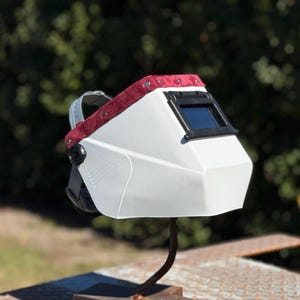 May include: A white welding helmet with a dark viewing lens and a red suede-like head support. The helmet is mounted on a metal stand, showcasing its design and features. The background is blurred, with green foliage.