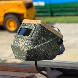 Op de afbeelding: Een lashelm met camouflagepatroon en een donkere kijklens. De helm heeft een leren bovenkant in beige en een zwarte afstelknop. De achtergrond omvat een gele bouwmachine en een groene structuur.