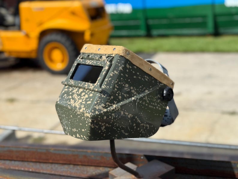 Op de afbeelding: Een lashelm met camouflagepatroon en een donkere kijklens. De helm heeft een leren bovenkant in beige en een zwarte afstelknop. De achtergrond omvat een gele bouwmachine en een groene structuur.
