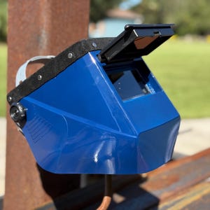 Op de afbeelding: Een blauwe lashelm met een zwart vizier en een zwarte bovenkant. De helm is iets omhoog gekanteld, waardoor de binnenkant zichtbaar is. De achtergrond is wazig, met een roestige metalen paal en groen gras.