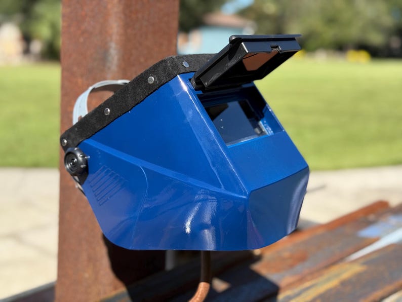 Op de afbeelding: Een blauwe lashelm met een zwart vizier en een zwarte bovenkant. De helm is iets omhoog gekanteld, waardoor de binnenkant zichtbaar is. De achtergrond is wazig, met een roestige metalen paal en groen gras.
