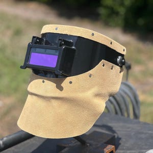 May include: A welding helmet featuring a black plastic face shield with a rectangular purple lens and a tan suede leather neck guard attached with metal rivets. The protective gear includes a side adjustment knob and a structured frame design.