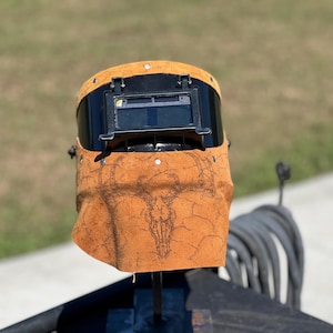 May include: A welding helmet with a black visor and a tan leather bib. The leather bib features a detailed graphic of a bull skull. The helmet is designed to protect the eyes and face during welding.