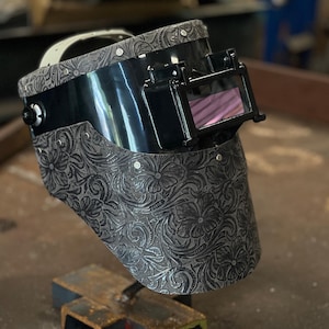 Puede incluir: Una careta de soldar con una sección superior negra brillante y una sección inferior gris grabada con un detallado patrón floral. El casco incluye un marco de lente rectangular abatible, una correa ajustable y remaches plateados.
