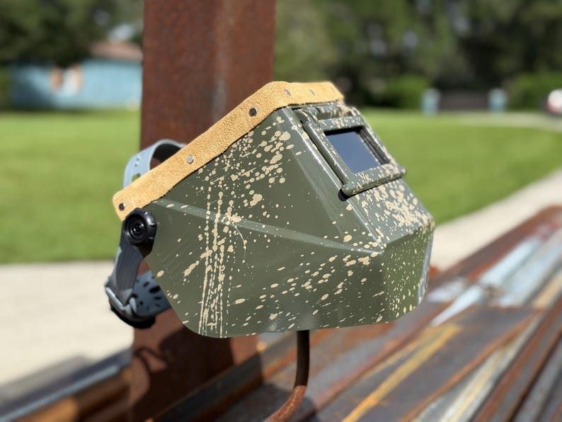 Op de afbeelding: Een olijfgroene lashelm met een spetterverf ontwerp. De helm heeft een donker kijkvenster en een bruin lederen rand langs de bovenrand. Een zwarte verstelbare hoofdband is bevestigd.