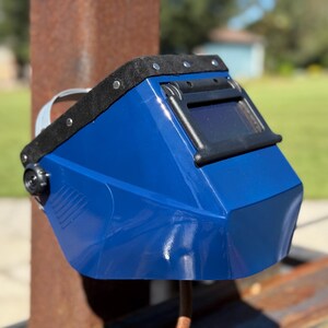 Op de afbeelding: Een blauwe lashelm met een zwart vizier en een zwarte gewatteerde hoofdsteun. De helm heeft een glanzende afwerking en een donkere lens. De helm is ontworpen om de ogen en het gezicht te beschermen tijdens het lassen.