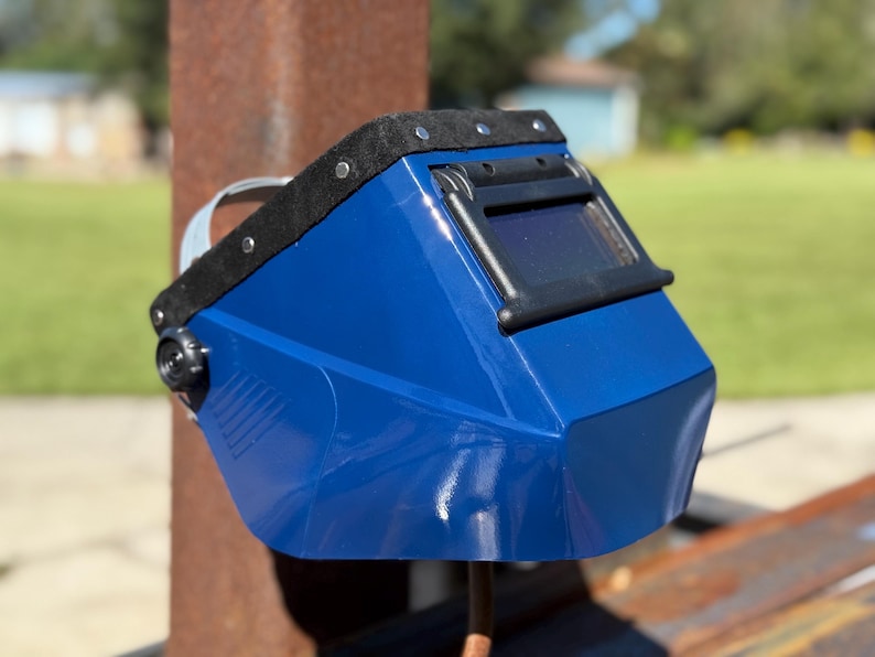 Op de afbeelding: Een blauwe lashelm met een zwart vizier en een zwarte gewatteerde hoofdsteun. De helm heeft een glanzende afwerking en een donkere lens. De helm is ontworpen om de ogen en het gezicht te beschermen tijdens het lassen.