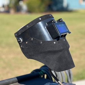 May include: A black welding helmet with a blue-tinted lens and a black suede-like bib. The helmet is secured with rivets and has a padded head support. The background is a blurred outdoor scene.