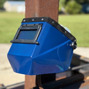 Op de afbeelding: Een blauwe lashelm met een zwart frame rond het kijkvenster. De helm heeft een zwarte, gewatteerde hoofdsteun en is gemonteerd op een metalen constructie. De achtergrond is wazig en toont groen gras en een gebouw.