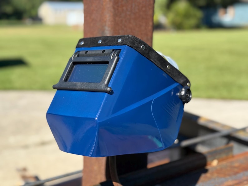Op de afbeelding: Een blauwe lashelm met een zwart frame rond het kijkvenster. De helm heeft een zwarte, gewatteerde hoofdsteun en is gemonteerd op een metalen constructie. De achtergrond is wazig en toont groen gras en een gebouw.