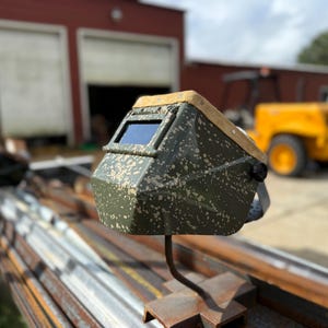 Op de afbeelding: Een groen en bruin gespikkelde lashelm met een donkere lens. De helm is gemonteerd op een metalen standaard. De achtergrond omvat een rood gebouw en een geel voertuig.