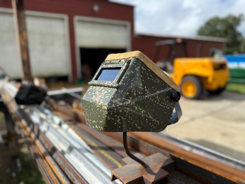 Op de afbeelding: Een groen en bruin gespikkelde lashelm met een donkere lens. De helm is gemonteerd op een metalen standaard. De achtergrond omvat een rood gebouw en een geel voertuig.