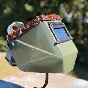 Puede incluir: Un casco de soldadura verde oliva con una lente oscura y una parte superior decorativa marrón texturizada. El casco tiene un marco negro alrededor de la lente y una correa para la cabeza gris. El casco está montado en un soporte de metal curvado.