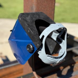 Op de afbeelding: Een blauw-zwarte lashelm met een grijs gelaatsscherm. De helm is gemonteerd op een roestige metalen constructie, met een groene achtergrond. De helm heeft een zwart interieur en een grijs gelaatsscherm.