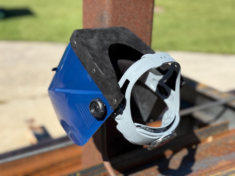 Op de afbeelding: Een blauw-zwarte lashelm met een grijs gelaatsscherm. De helm is gemonteerd op een roestige metalen constructie, met een groene achtergrond. De helm heeft een zwart interieur en een grijs gelaatsscherm.