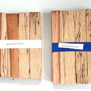 May include: Two sets of spalted red ash wood, each set containing five pieces of wood. The wood is light brown with dark brown streaks. The sets are labeled "Spalted Red Ash #21" and "Spalted Red Ash #22".