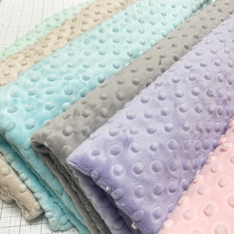 May include: A stack of six fabric swatches in various colors, including light pink, lavender, gray, light blue, beige, and green. The fabric has a textured, raised dot pattern.