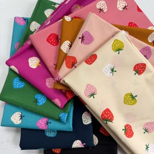 May include: A collection of folded fabric squares in various colors, including pink, green, and blue. Each fabric features a repeating pattern of strawberries in red, pink, yellow, and white. The strawberries have green stems and are scattered across the fabric.