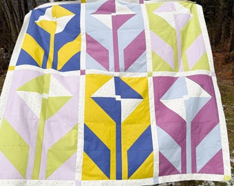 May include: A quilt featuring a grid of six large floral blocks. Each block displays a stylized flower in shades of yellow, blue, purple, and light green. The blocks are separated by white sashing with a subtle pattern of small dots.
