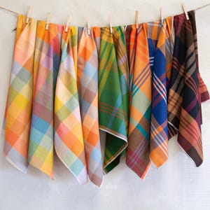 May include: A collection of colorful plaid fabric cloths hanging from a clothesline. The cloths feature a variety of colors, including orange, green, blue, and purple. The cloths are made of a soft material and are likely used as kitchen towels or decorative items.