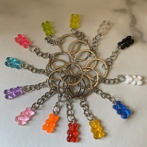 May include: A set of 12 colourful gummy bear keyrings with silver key rings and chains. The keyrings are arranged in a circle on a white surface.