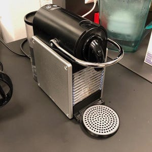 May include: A black and silver De'Longhi coffee machine with a sleek, rectangular body, a chrome lever, a textured side panel, and a circular drip tray. The unit features a clear water reservoir at the back and a top control panel with buttons.